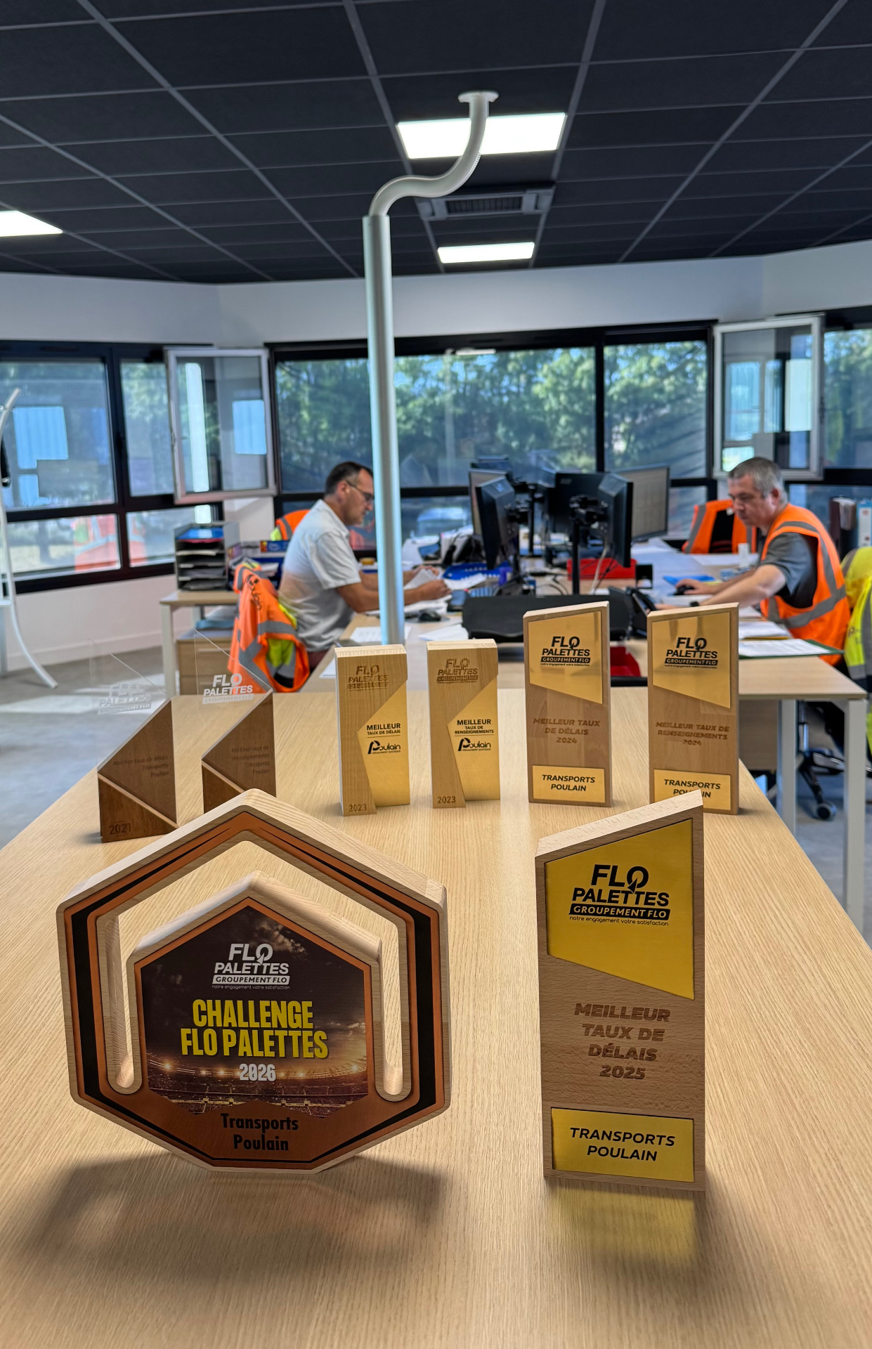 Deux nouveaux trophées récompensent l'engagement quotidien des Transports Poulain Deux trophées FLO Palettes posés sur un bureau, avec les exploitants des Transports Poulain en arrière-plan