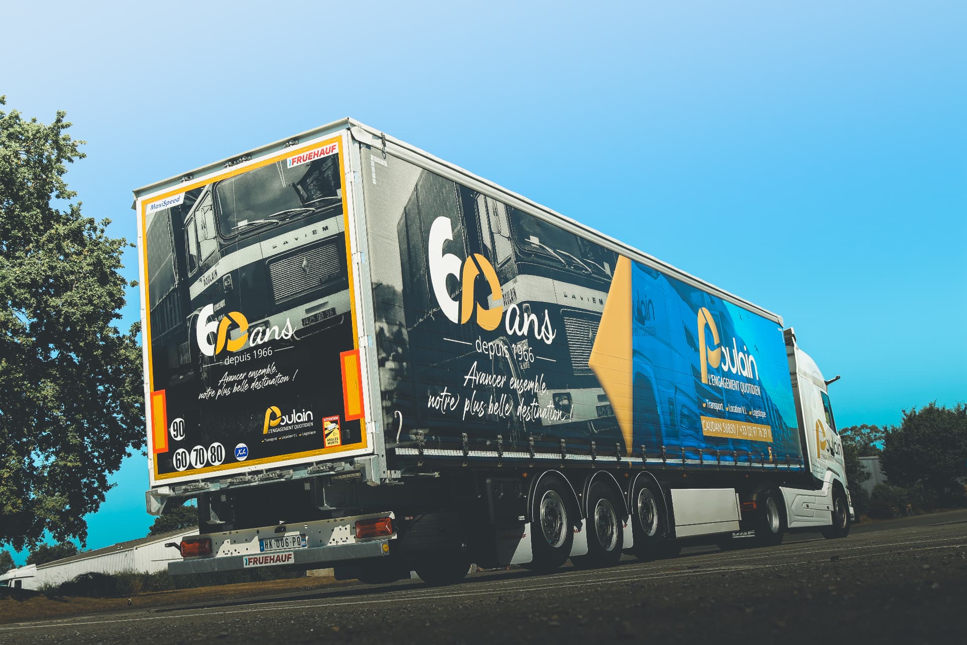 Contre-plongée d'un camion Poulain avec remorque décorée 60 ans noir, blanc et jaune, sous un ciel bleu dégagé